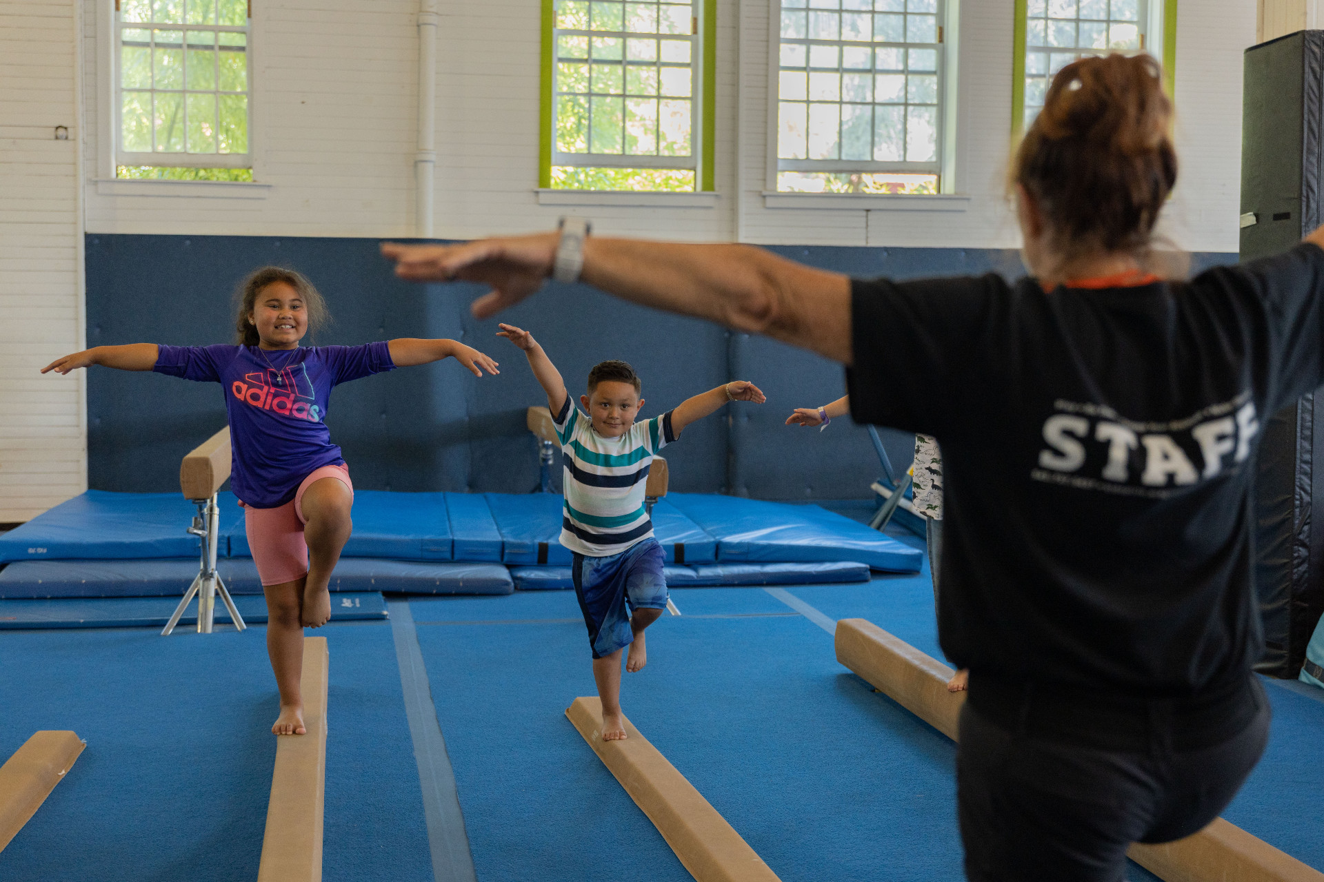 2 children balancing on a gymnastics beam, mimicking a woman demonstrating