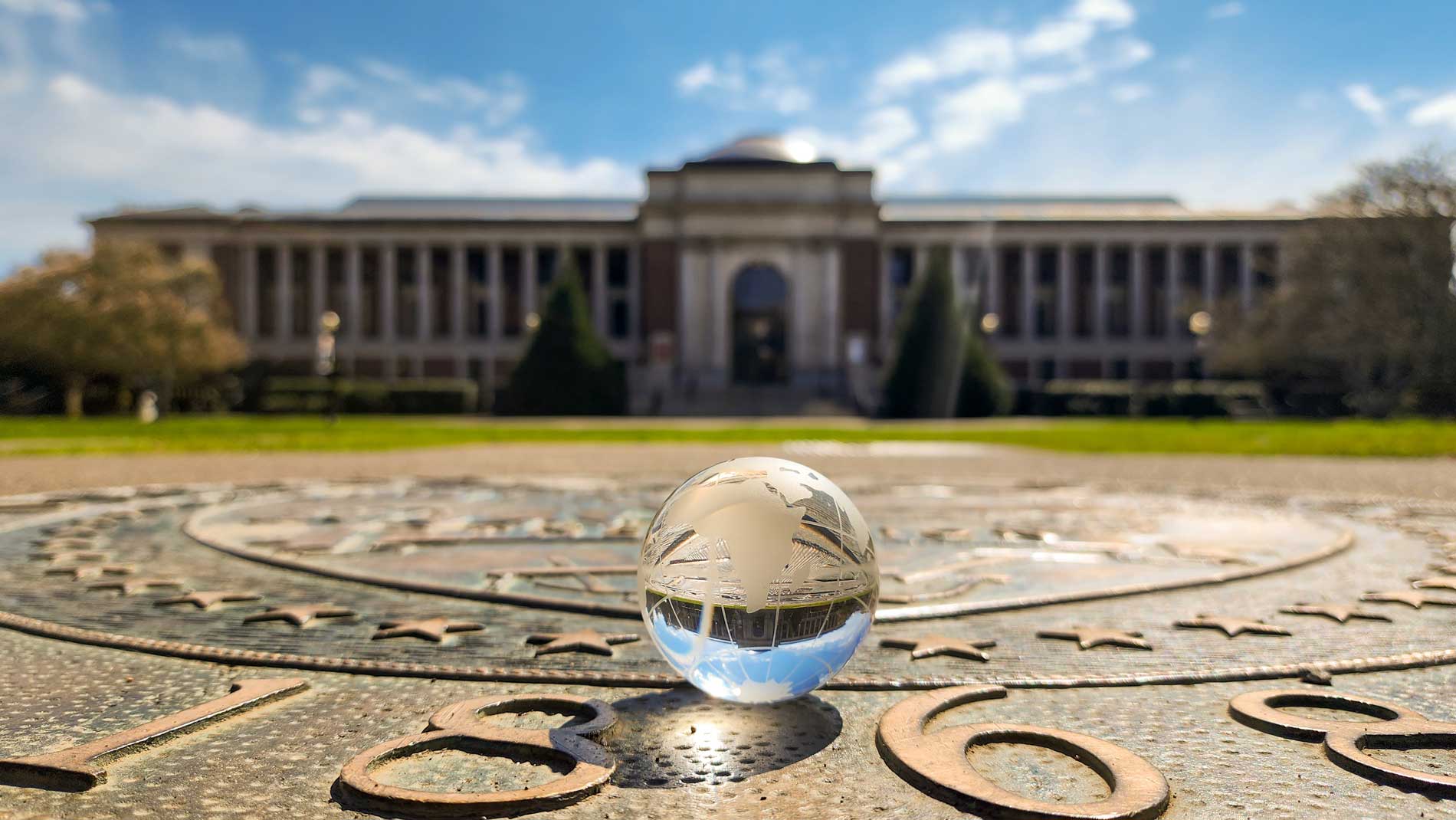 a glass globe sitting on a plaque in the ground that reads 1868 with the Memorial Union building in the background