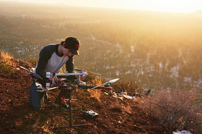 person squatting next to a drone on the side of a hill with scrub brush at sunset