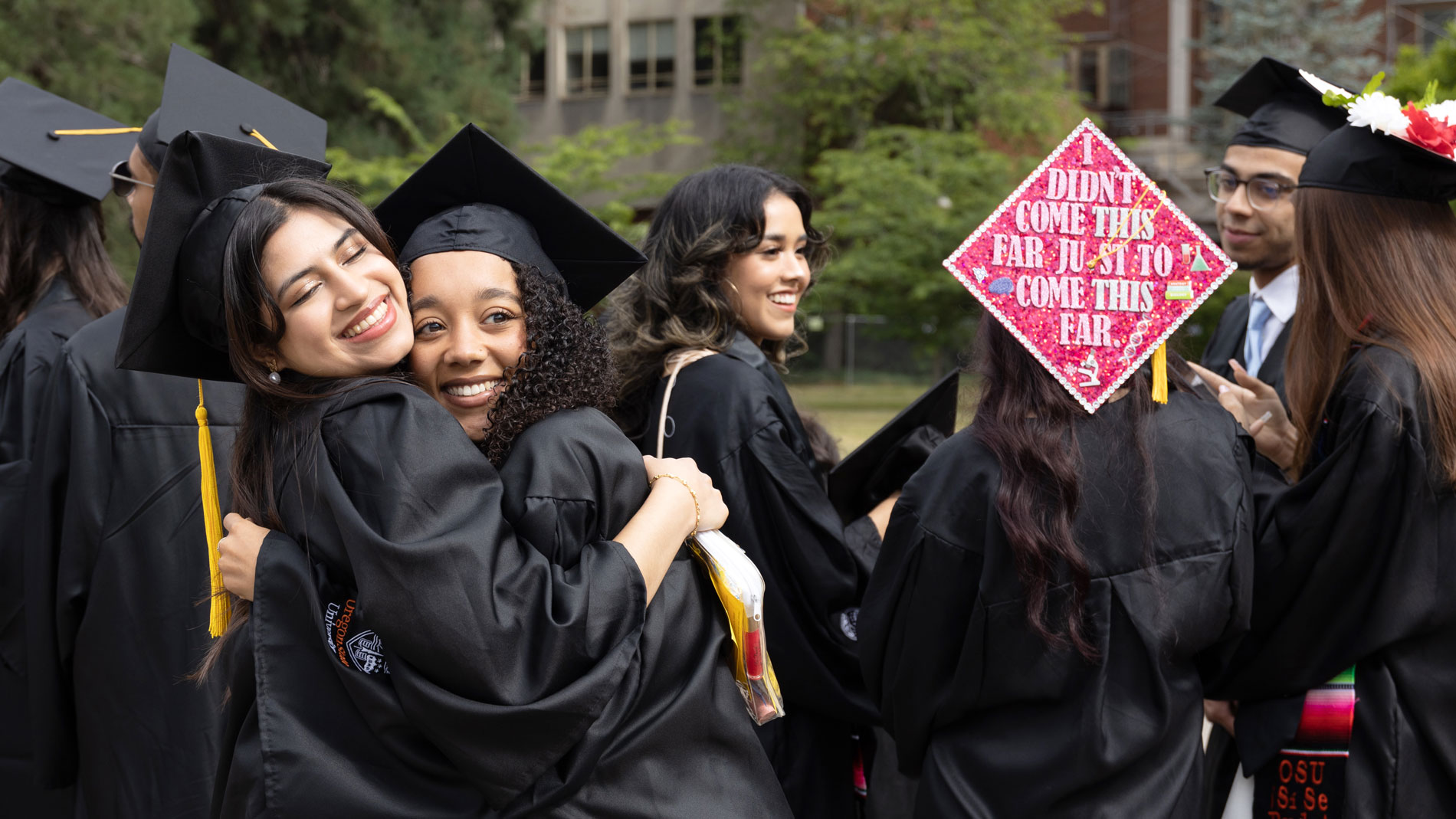  group of graduates in caps and gowns hugging and smiling at a graduation ceremony.